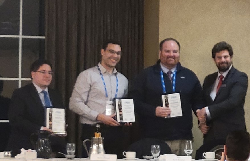 Four men in a row with three holding plaques and one man awkwardly shaking the hand of another man