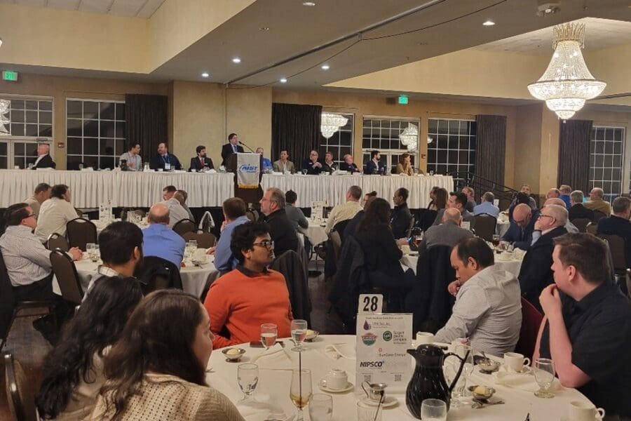 Several round tables with people sitting in chairs at them. Long table in the front of the room with man at podium.