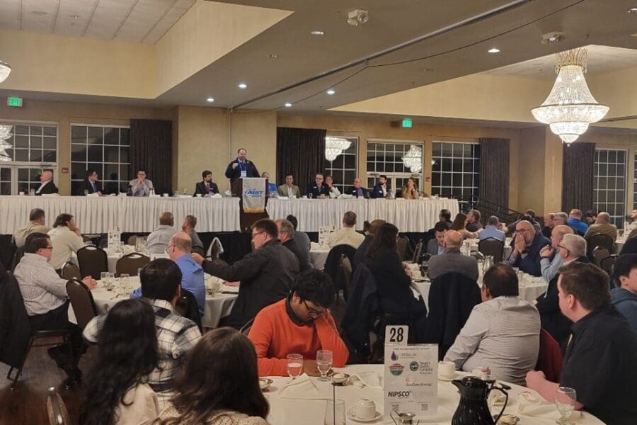 Several round tables with people sitting in chairs at them. Long table in the front of the room with man at podium.