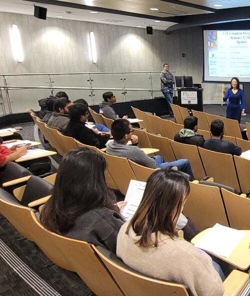 Woman standing in front of screen in auditorium with several students seated in rows of chairs