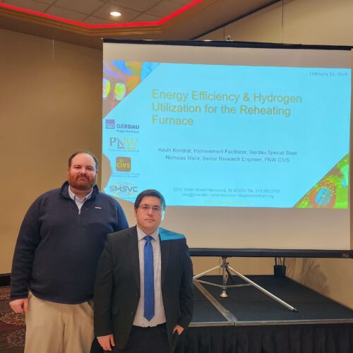 Two individuals in business attire standing in front of a screen with a PowerPoint titled Energy Efficiency & Hydrogen Utilization for the Reheating Furnace
