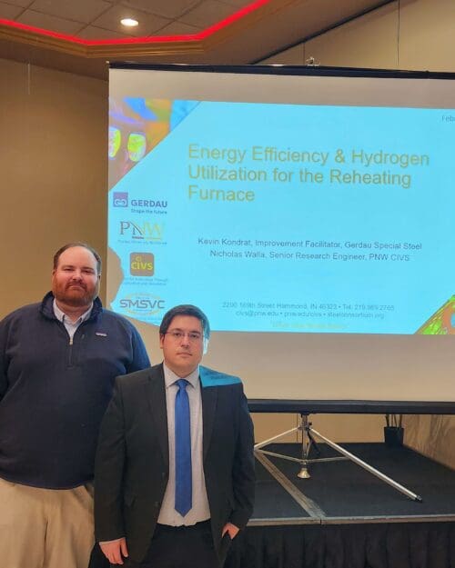 Two individuals in business attire standing in front of a screen with a PowerPoint titled Energy Efficiency & Hydrogen Utilization for the Reheating Furnace