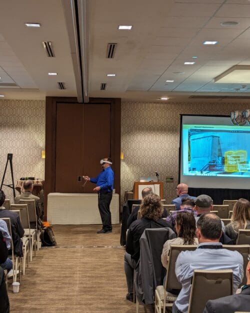 Conference rooms with people in rows of chairs facing two large screens. Jack Moreland wearing a VR headset and manipulating hand controls at the front of the room.