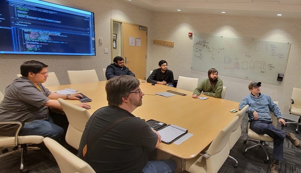 A group of individuals sit around a conference table looking at a large screen with various technical data on it