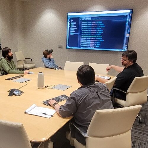 A group of individuals sitting around a conference table looking at a large screen displaying various technical data