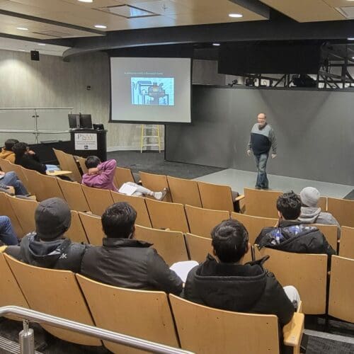 Man standing in front of screen in auditorium with several students seated in rows of chairs