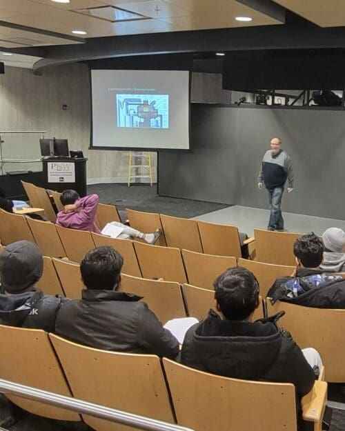 Man standing in front of screen in auditorium with several students seated in rows of chairs