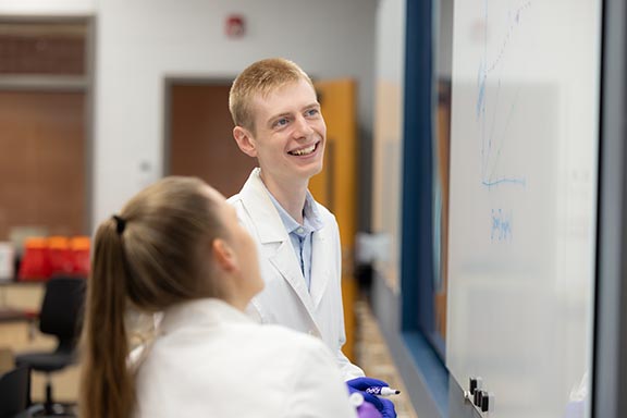 Two PNW students writing on the whiteboard in a classroom while talking with one another