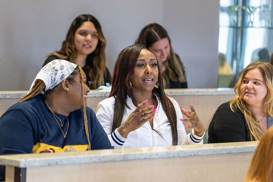 Students sit in class. They are sitting in two rows of tables. All the students but one are looking at the student sitting in the middle of the front row.