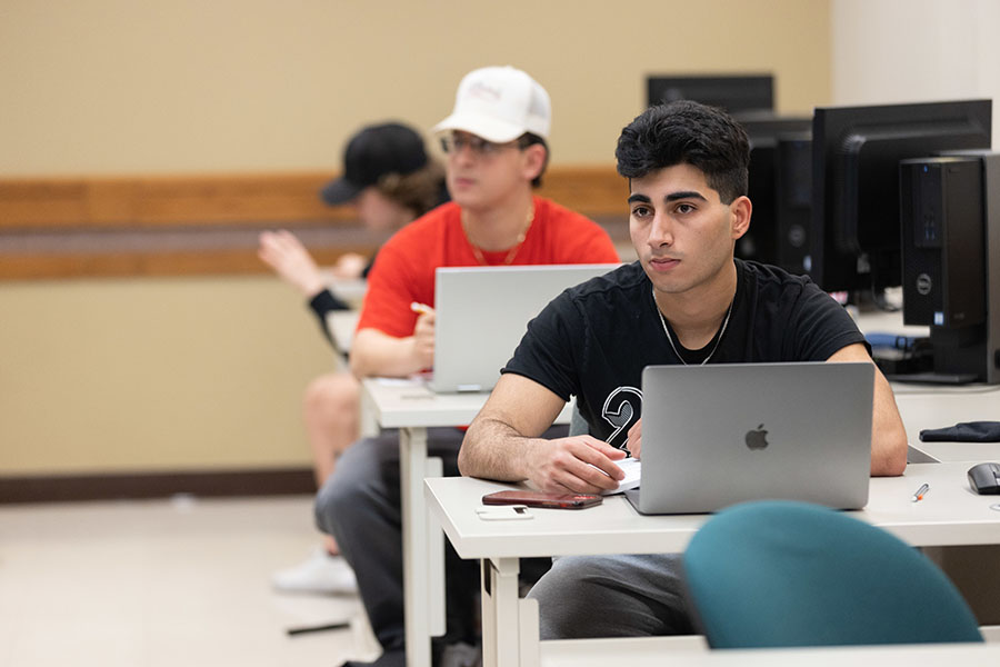 Students sit in a row during class. There are three students pictured. All three students have laptops in front of them.