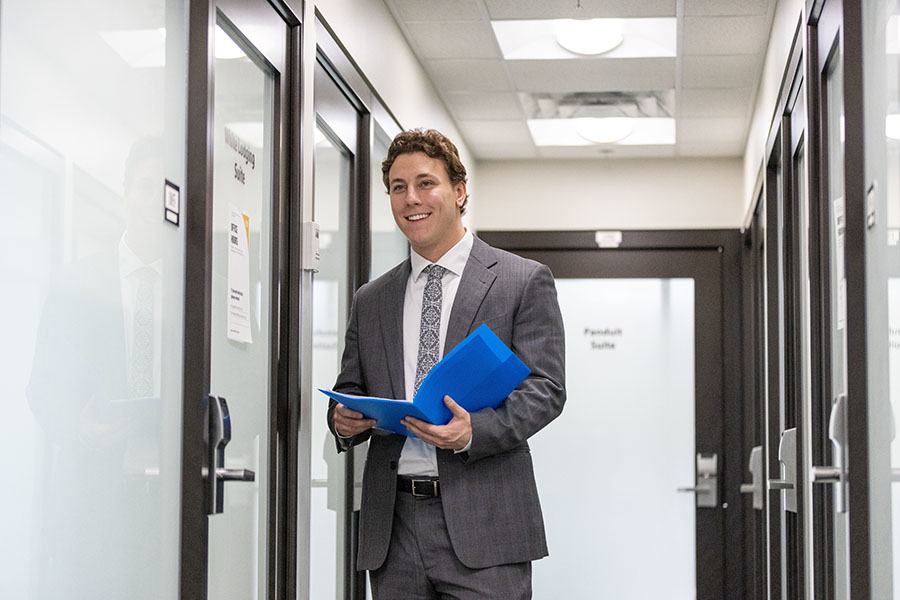 Student stands in a hallway in a suit. He is holding a blue folder