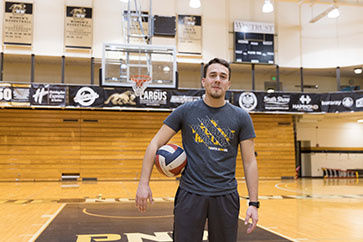 A student stands on a volleyball court with a volleyball under their arm.