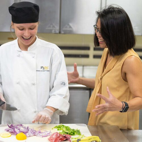 A student stands with an HTM instructor in the kitchen. The student is chopping food on a chopping board.