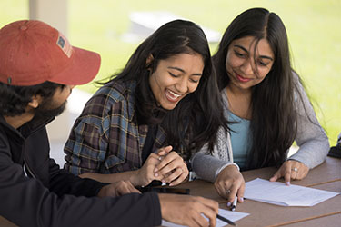 Three students sit together at a picnic table outside. There is a piece of paper on the table that they are looking at.