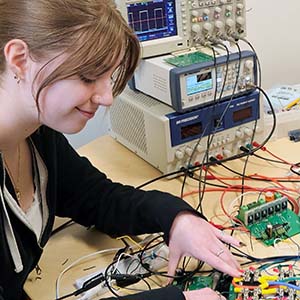 A woman works on a circuit board next to some computer equipment.