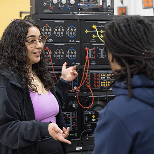 Two students work in an engineering lab. One student is facing the camera and gesturing toward the equipment. The other student is sitting, facing away from the camera.