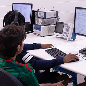 Two students sit at a computer and work together. One student has the keyboard and mouse in front of them, the other is watching.