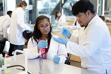 Two students in white lab coats work in a classroom lab space.