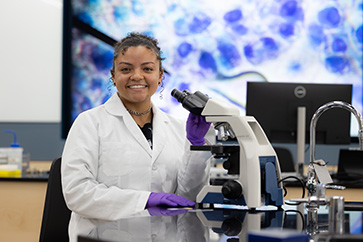 A student in a white lab coat sits at a microscope.