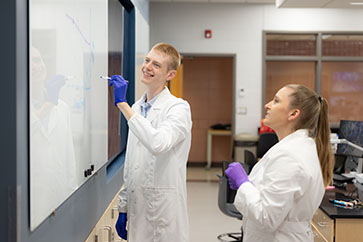 A student writes on a board with a dry erase marker. A professor is standing behind him.