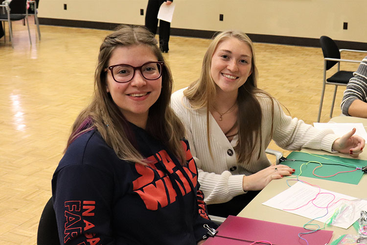 Two students sit together at a table and smile at the camera