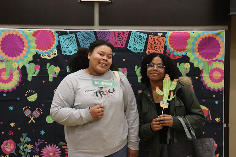 Two students hold up props and pose in front of a backdrop