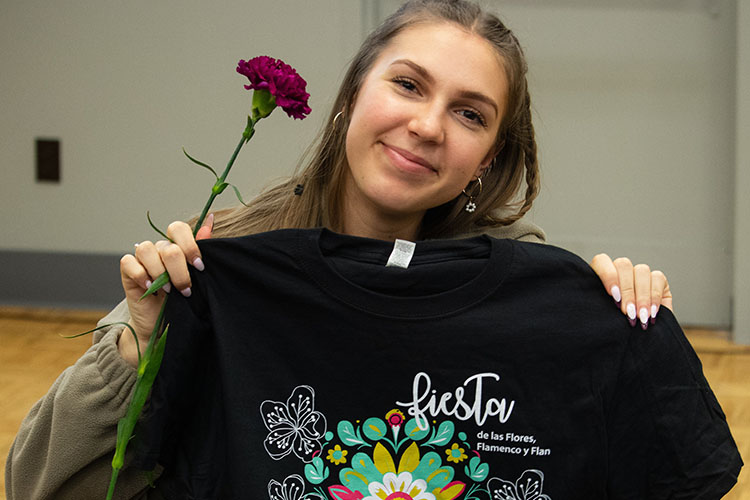 A student smiles and holds up a carnation and a Fiesta de las Flores, Flamenco y Flan t-shirt