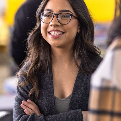 A student in a light gray shirt and dark gray cardigan stands with their arms folded. They are looking off to the left and smiling.