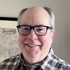 Headshot of Mike Lynn in a plaid shirt in front of a framed map.