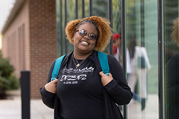 A student poses outside wearing a backpack. They are holding onto the straps of the backpack.