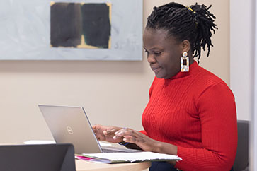 A student in a red sweater sits at a table and works on a laptop.