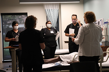 Four students grouped around a mannequin while the professor is teaching
