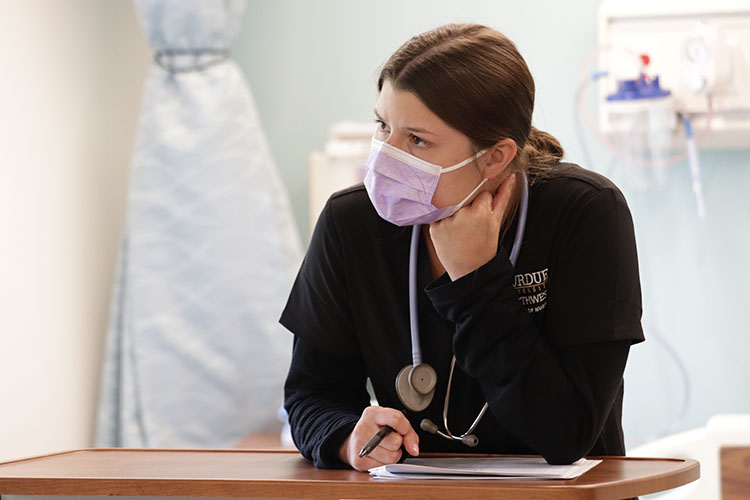 A nursing student in black scrubs and a mask writes notes in a lab.