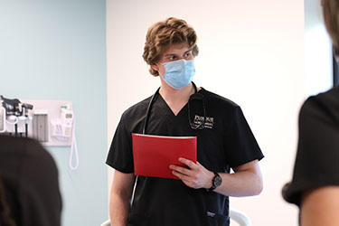 A nursing student in a mask holds up a folder and stands in a nursing lab