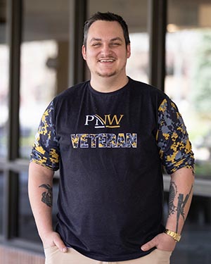 A man with military tattoos in a camouflage-style "PNW Veterans" shirt.