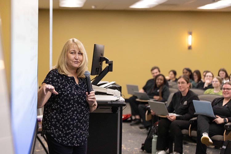 A lecturer with a microphone speaks to a classroom of nursing students.