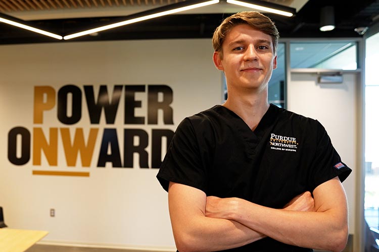 A student in PNW nursing gear stands with crossed arms in front of a Power Onward banner