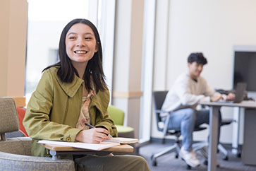 A student in a green jacket and floral shirt sits at a desk. Their hand is resting on an open notebook on the desk. They are holding a pen.