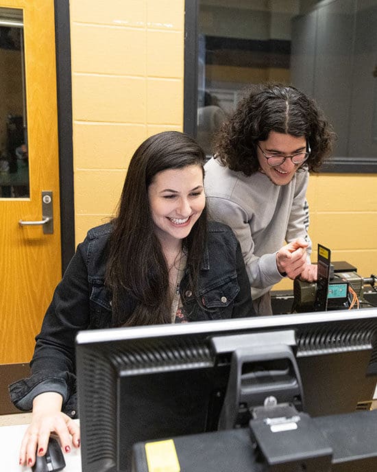 Two students look at a desktop computer. One student is standing and the other student is sitting.