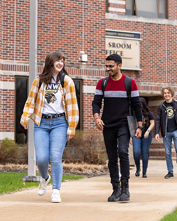 Two students walk together outside of the Classroom Office Building. There are more students on the sidewalk behind them