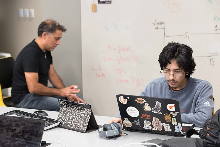 student working on their laptop with a professor writing on the whiteboard in the background