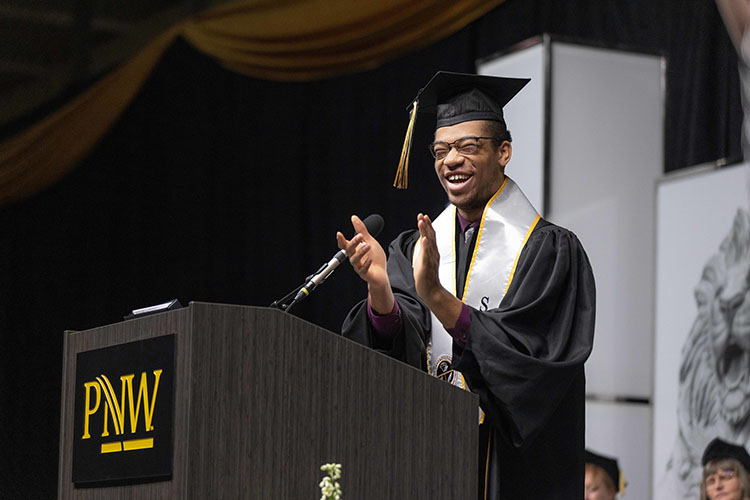 A young man in a cap and gown claps at a podium