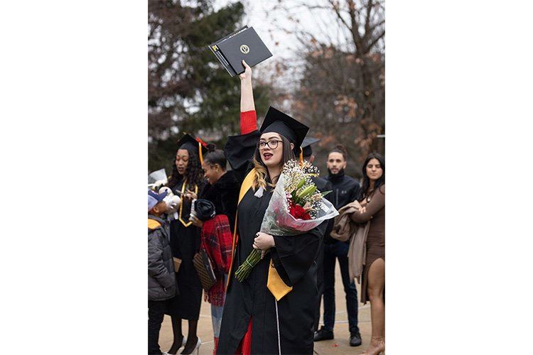 A PNW graduate with flowers holds her diploma high.
