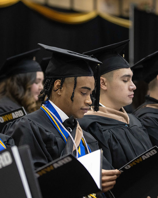 Students sit in rows during commencement. The photo focuses on two students who are looking at the commencement booklet they are holding. Everyone is wearing black commencement regalia.