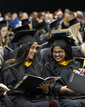 Two students sit together during commencement. They are looking at a commencement booklet