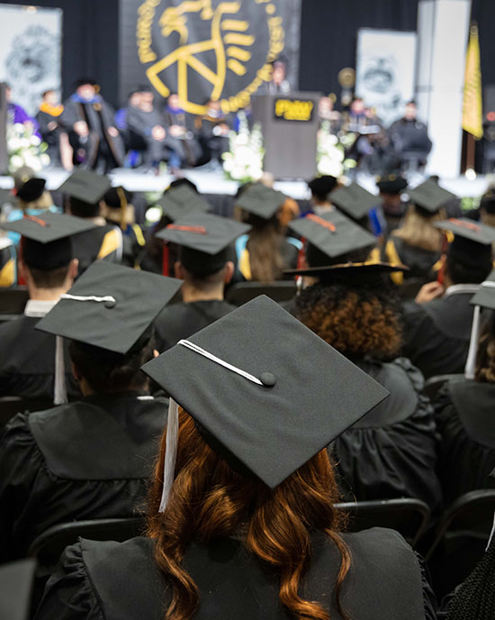 Rows of students facing the stage during an indoor commencement ceremony. Everyone is wearing black commencement regalia.