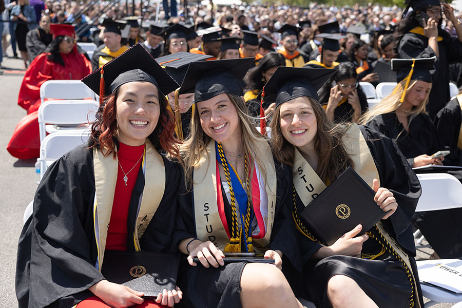 Three students lean together while sitting in the commencement crowd
