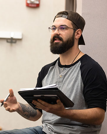 A student sits on a table in front of a class. They are holding papers in one hand and a pen in the other.