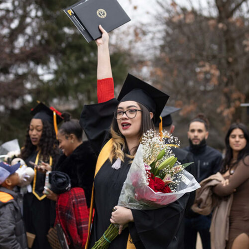 A PNW graduate with flowers holds her diploma high.