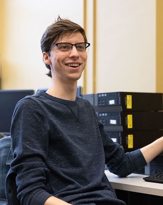 A student sits at a desk. Their elbow is propped on the desk and there is a stack of computers behind them.